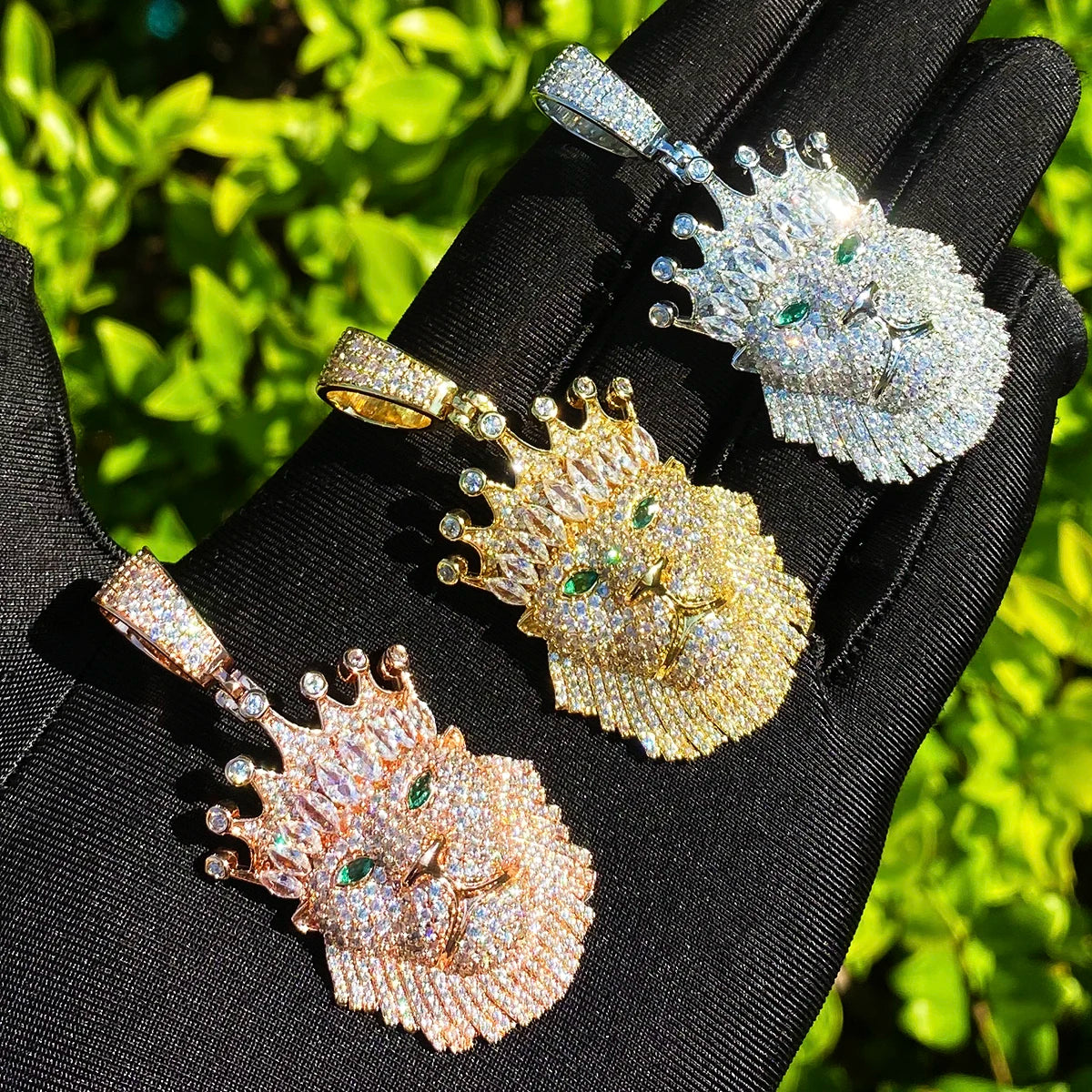 Four lion head pendants in gold, rose gold, and silver on a black glove with a green leafy background.