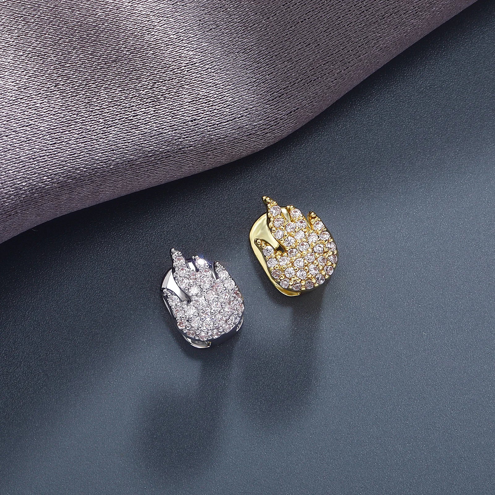 A pair of 18K gold diamond-studded fire emoji grillz on a grey surface.