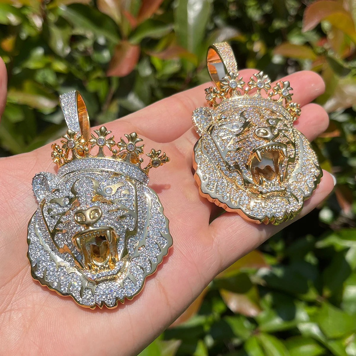 Two ornate tiger head pendants held in a hand with a green leafy background.