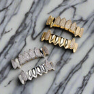 A set of gold and silver fang grillz, which are dental accessories designed to resemble sharp fangs, laid on a marble surface. The grillz are embellished with sparkling diamond-like stones.