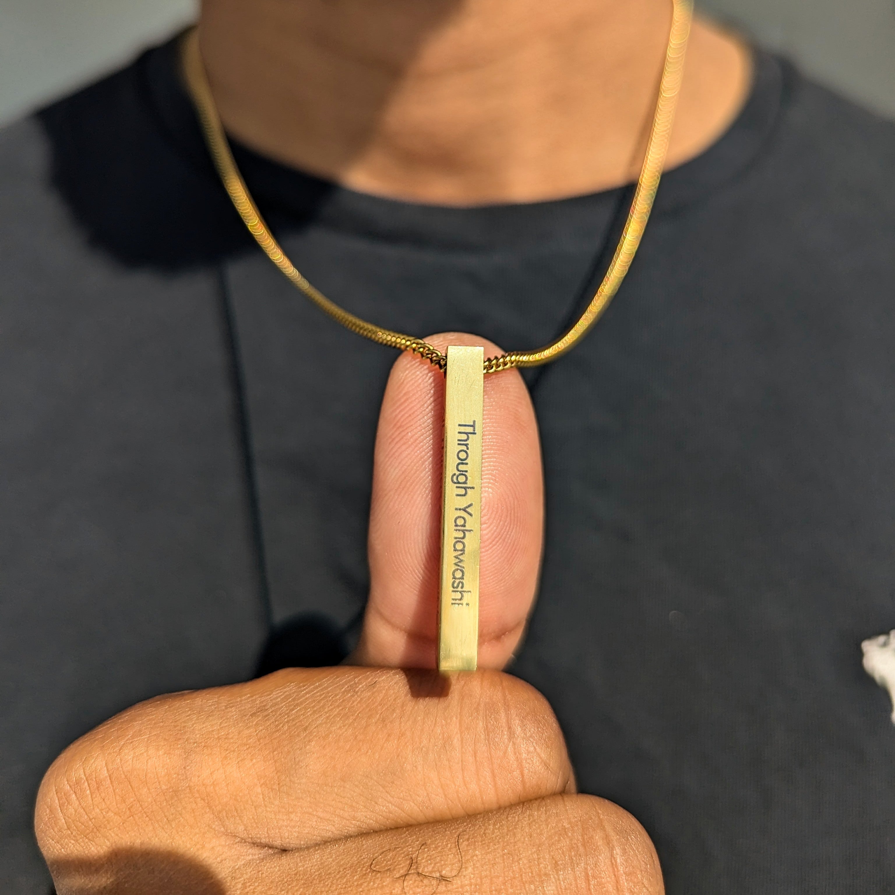 A person holding a custom 18k gold bar pendant on a chain, with engraving visible on the pendant.
